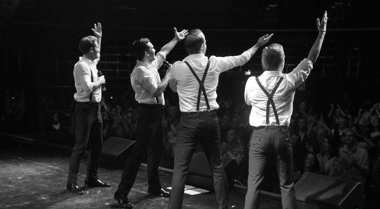 Four men in white shirts and grey trousers wearing dark suspenders look out over an audience, holding microphones. 