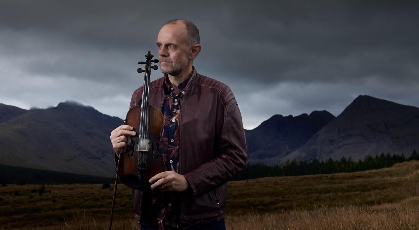 Duncan Chisholm stands holding his violin with the Black Cuillin mountains behind him