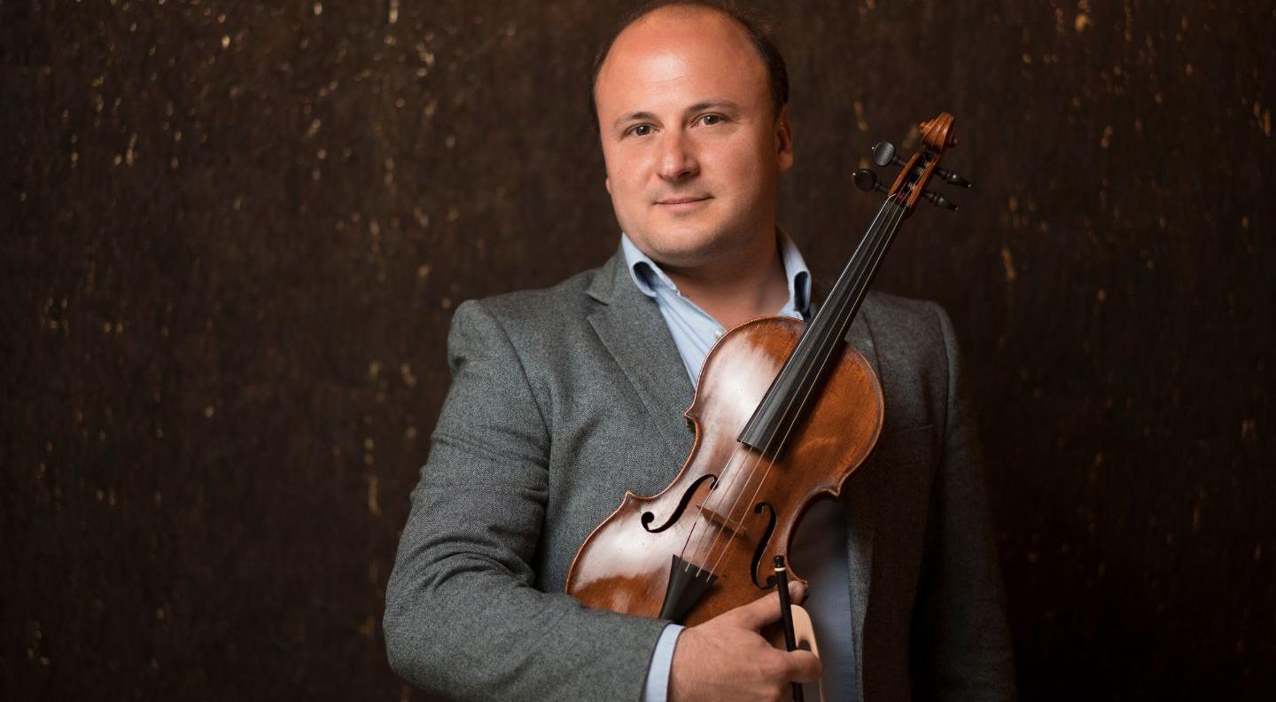 A white man wearing a grey suit stands against a dark background, holding a violin 