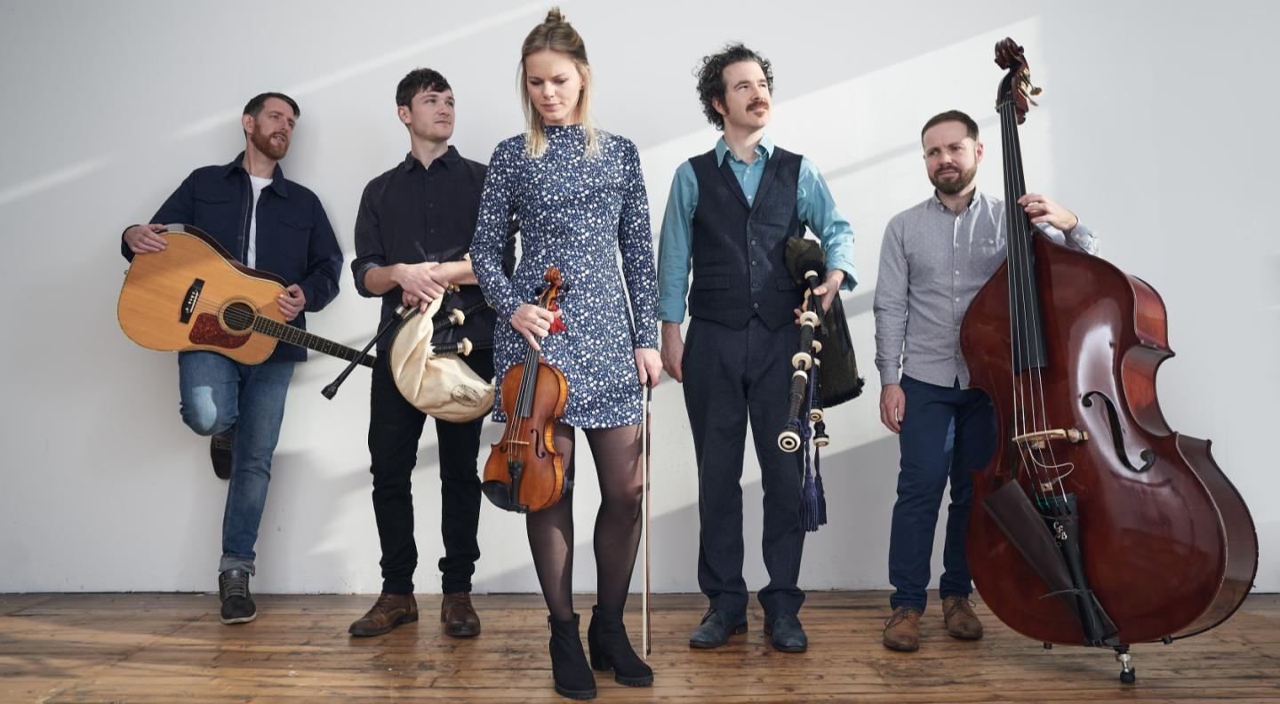 The 5 members of the band Breabach stand against a white wall, holding their instruments