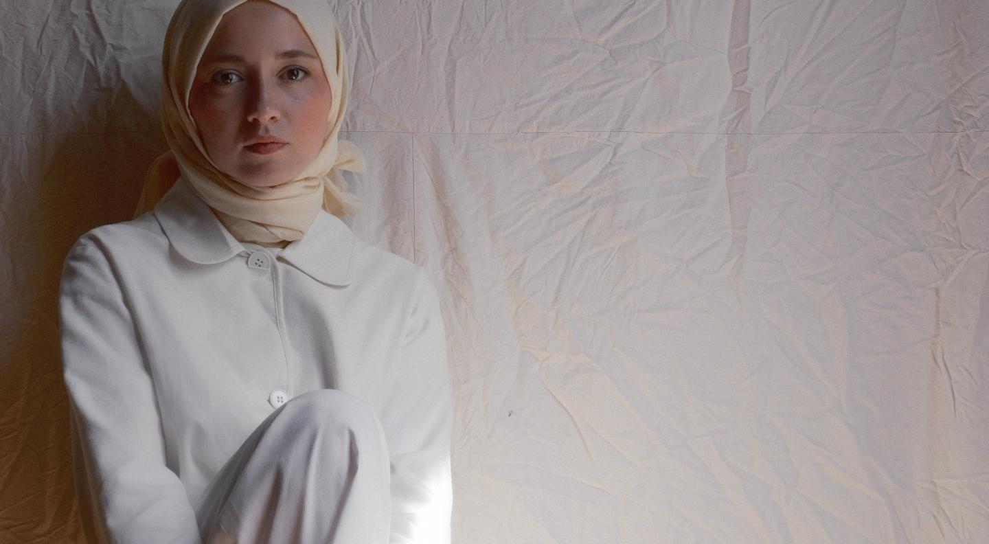 A young woman dressed all in white and wearing a light coloured headscarf leans against a light background with her knee slightly raised looking out at the viewer