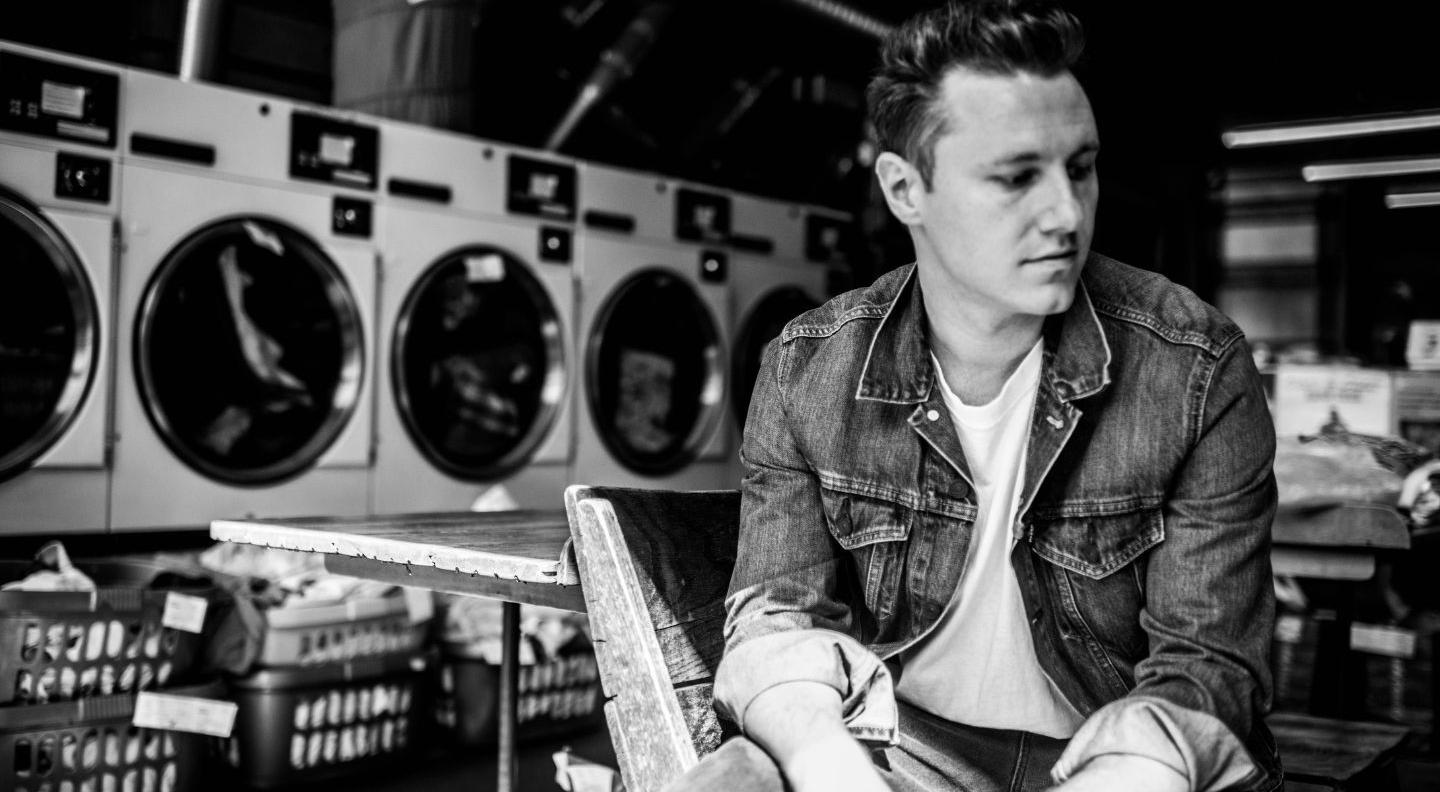 A black and white image of Callum, a white man wearing a denim jacket, sat in a room with lots of washing machines.