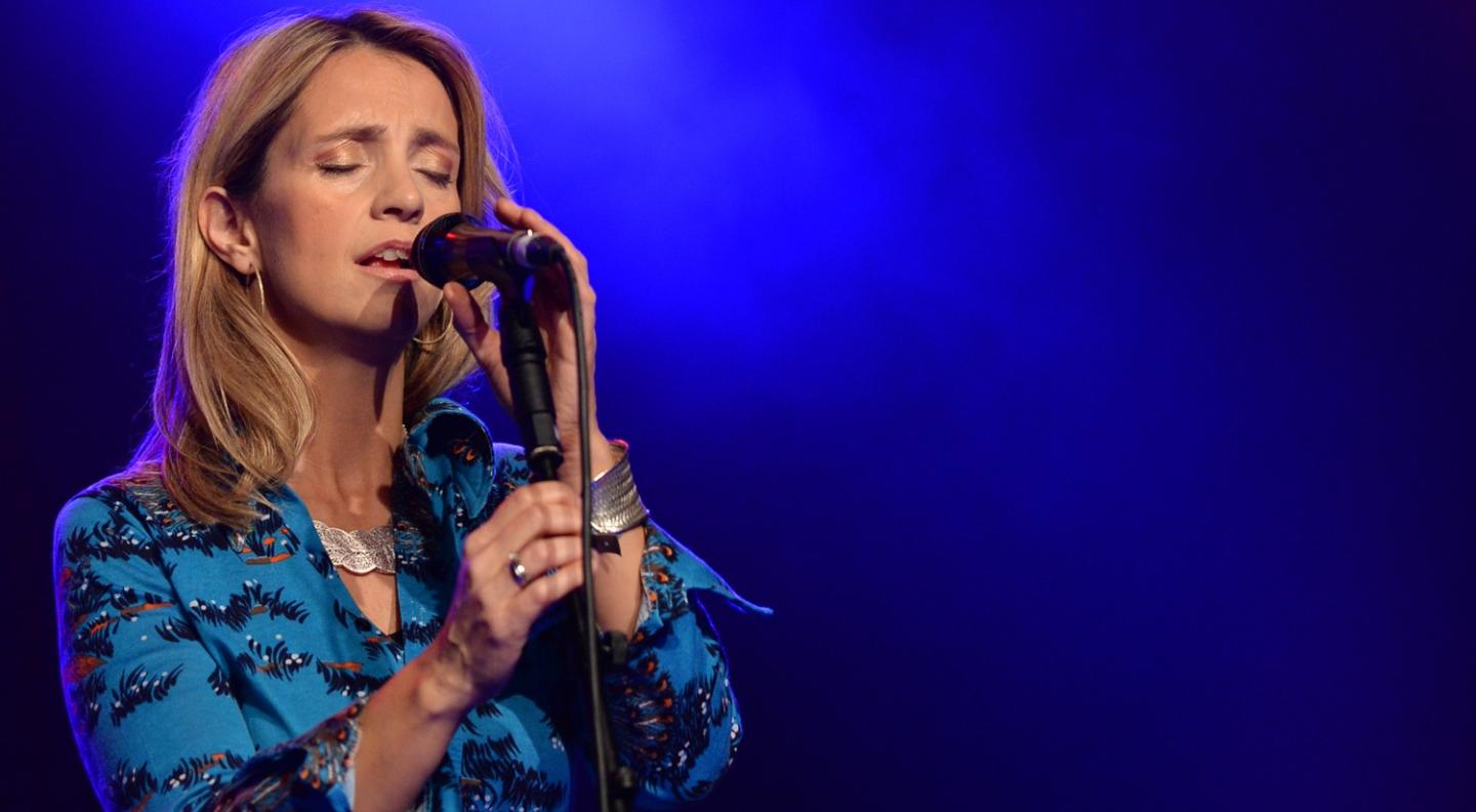 Cara Dillon wears a blue shirt with a black, white and orange pattern, and silver jewellery. She sings into a microphone stand with her eyes closed. There is a blue, smoky, lit background behind her.