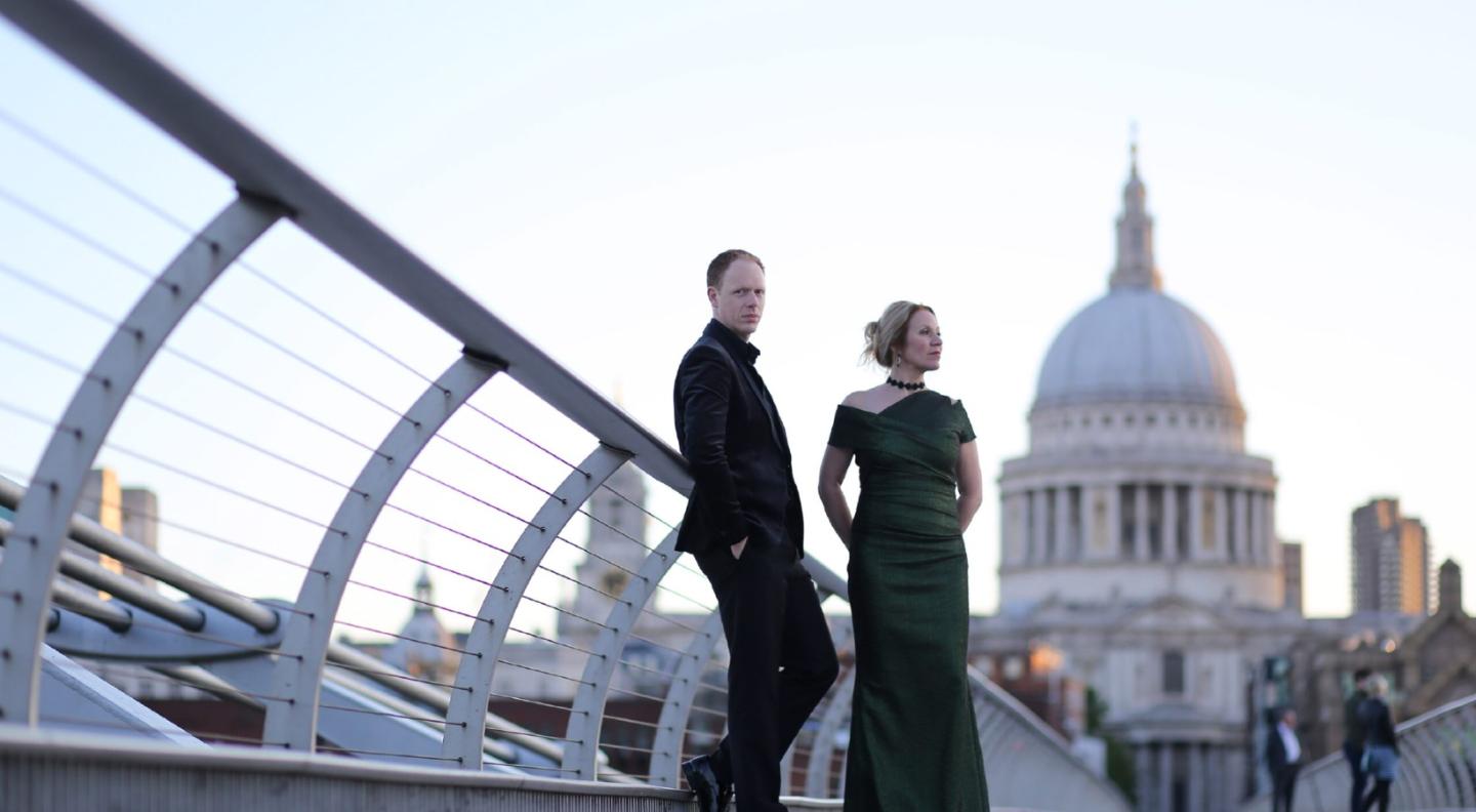 Carolyn Sampson and Joseph Middleton, a white woman and man stand in evening dress on the Millenium Bridge in London. St Paul's Cathedral is visble behind them