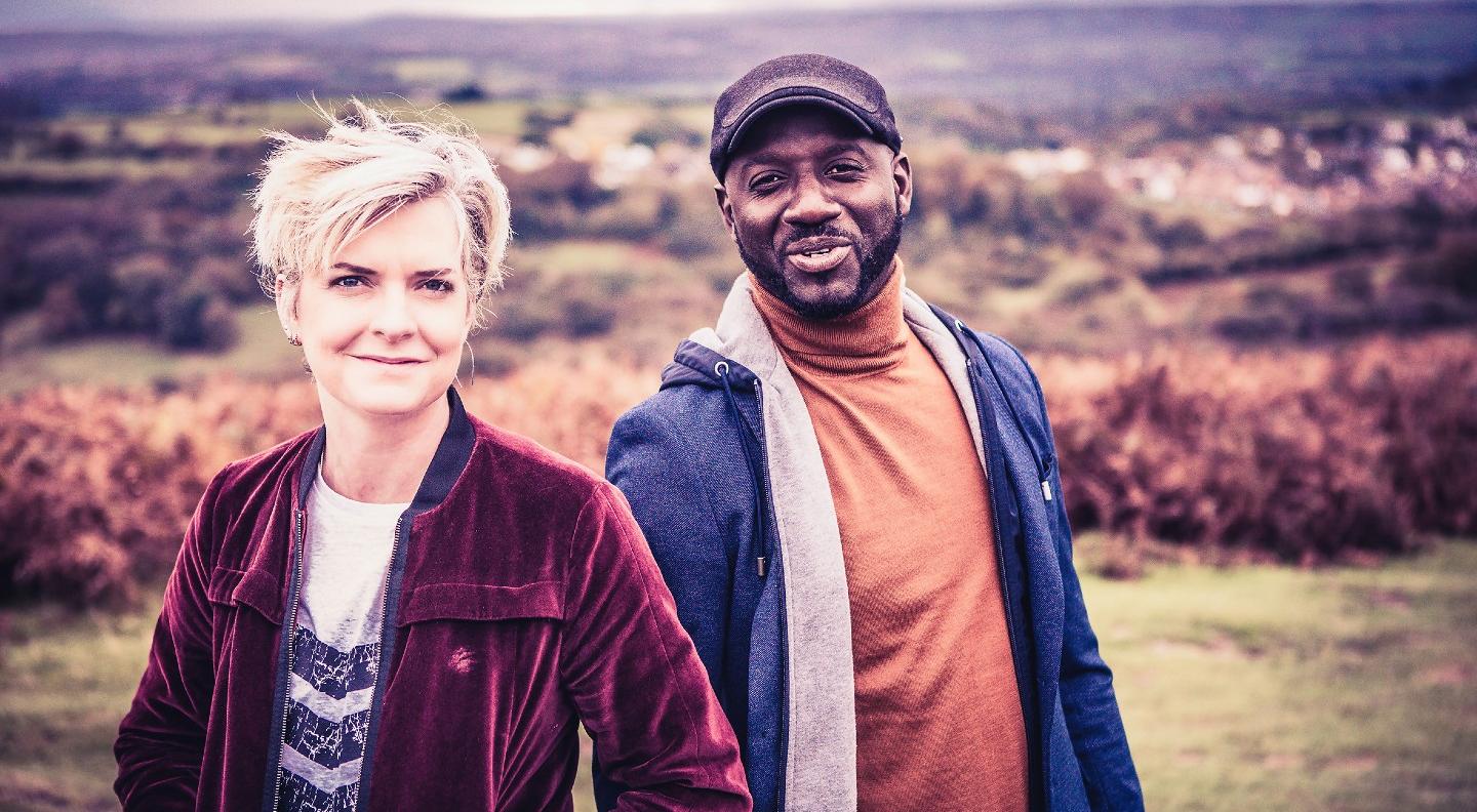 Catrin Finch and Seckou Keita stand in an outdoor countryside setting smiling at the camera