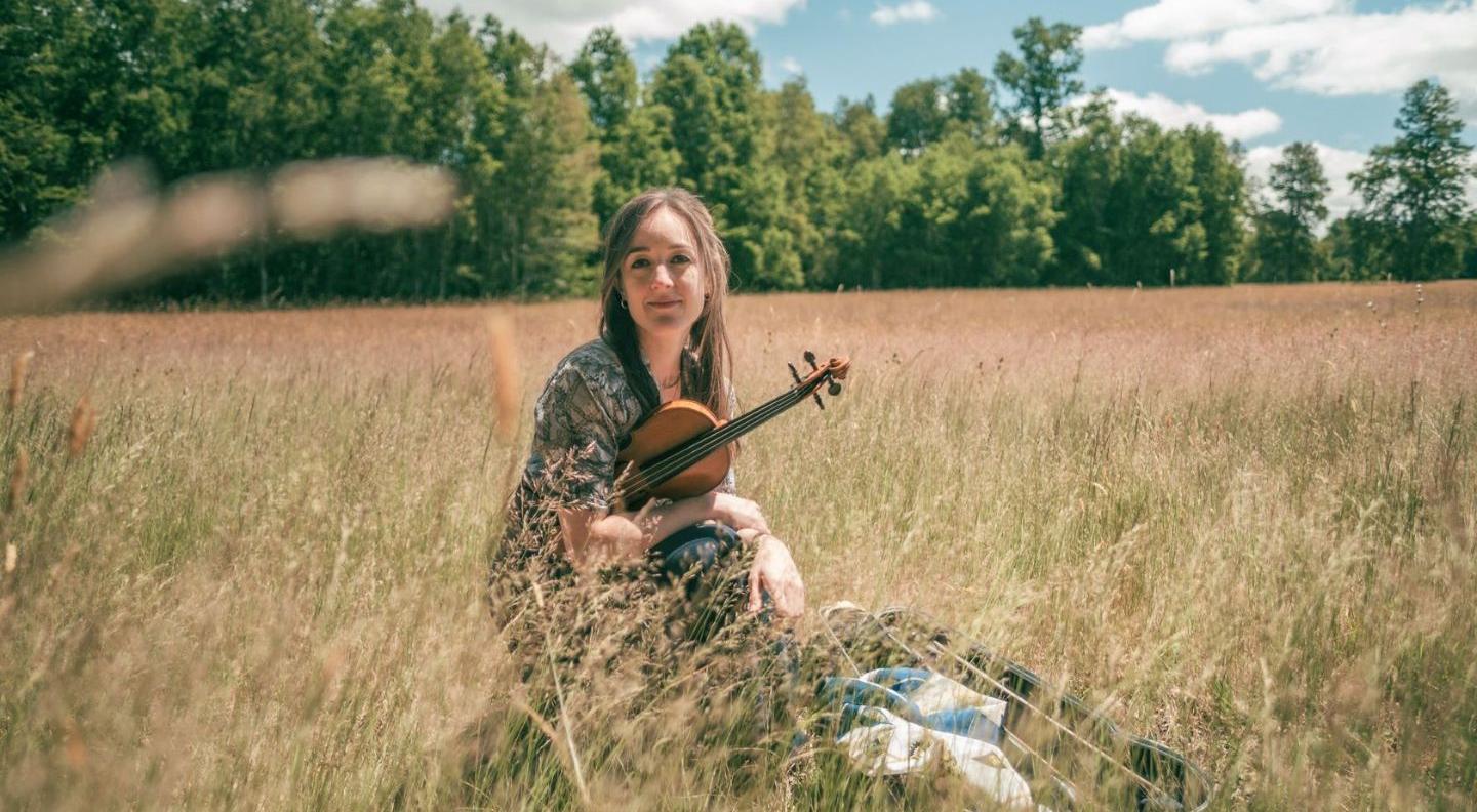 A white woman with long brown hair sits in the middle of a hay field with trees in the background, holding a violin