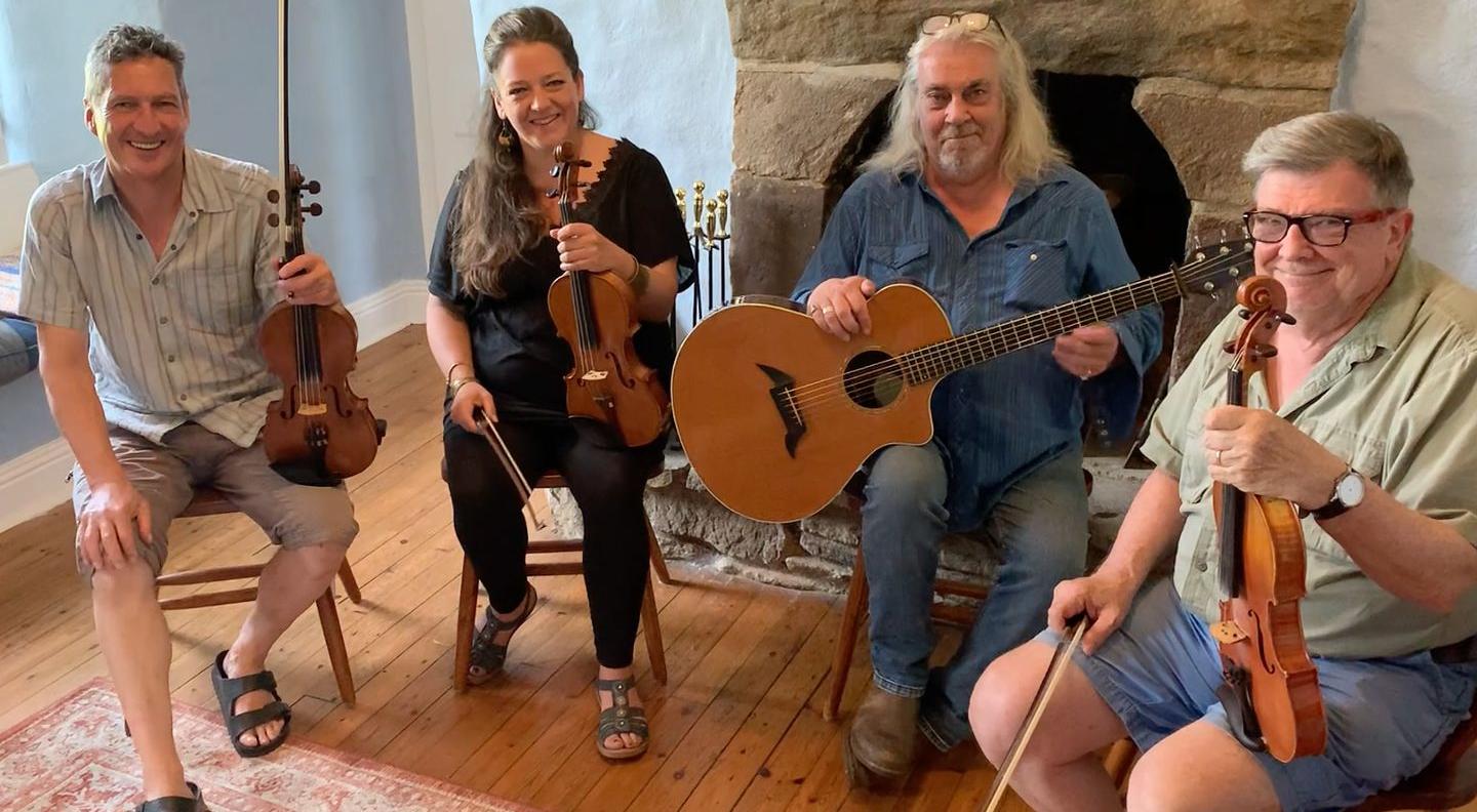 The Celtic Fiddle Festival musicians sit in a living room holding their instruments