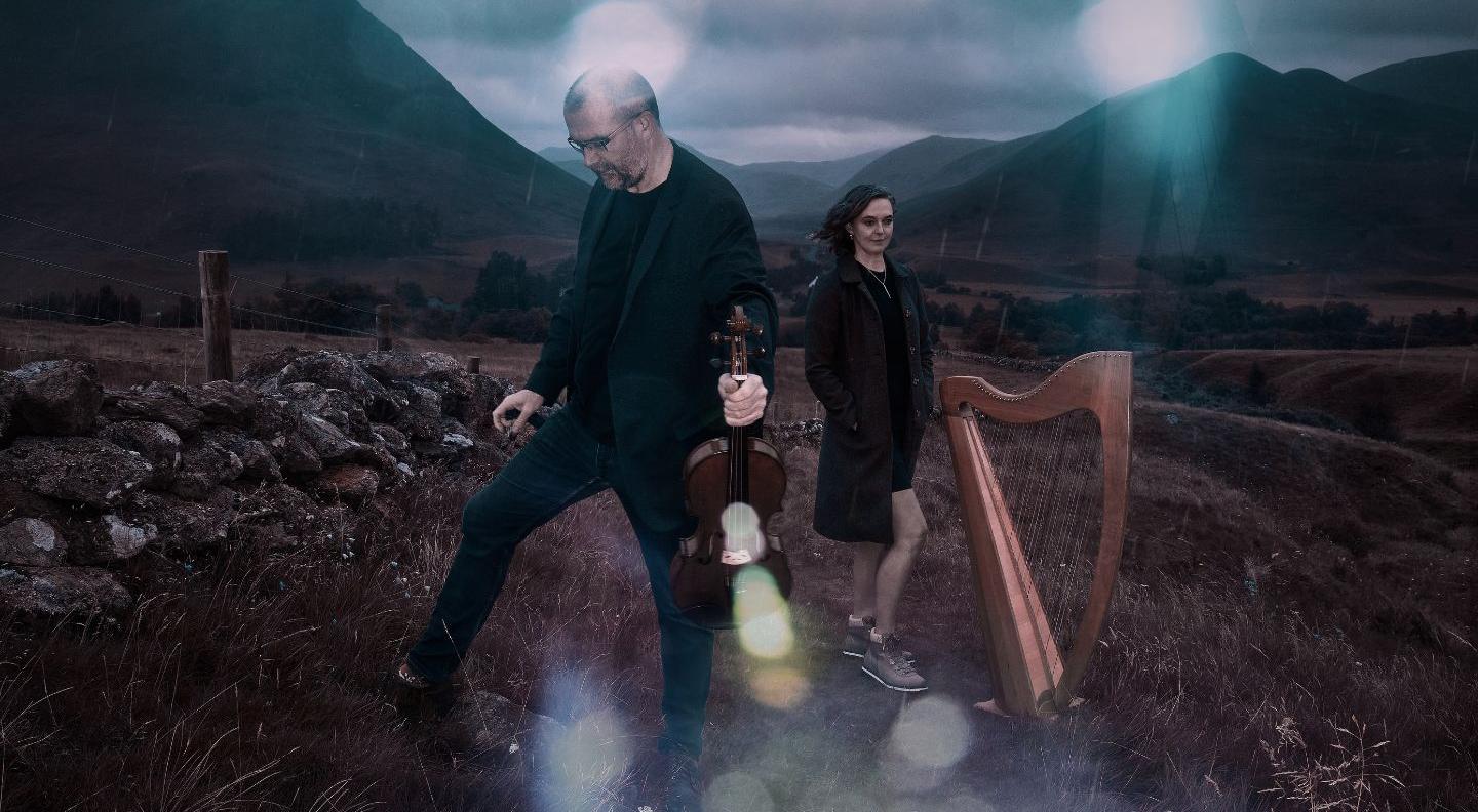 Chris and catriona in the Scottish Highlands, holding a violin and harp respectively