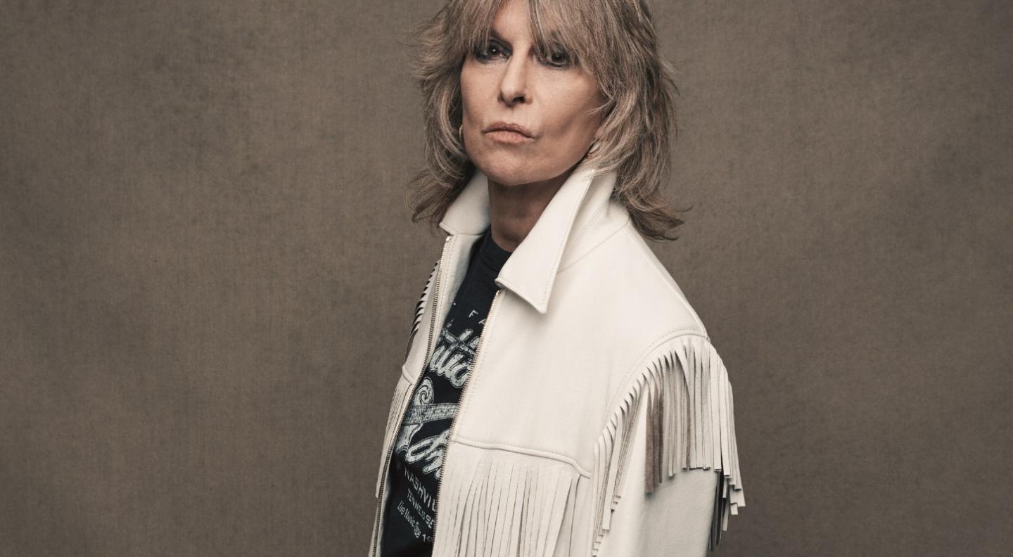 A mid shot of Chrissie Hynde standing side on against a plain grey/brown background. Chrissie wears a white fringed leather jacket with the collar up and a black t-shirt with partially visible, but unreadable white writing. She stares at the camera intently. 