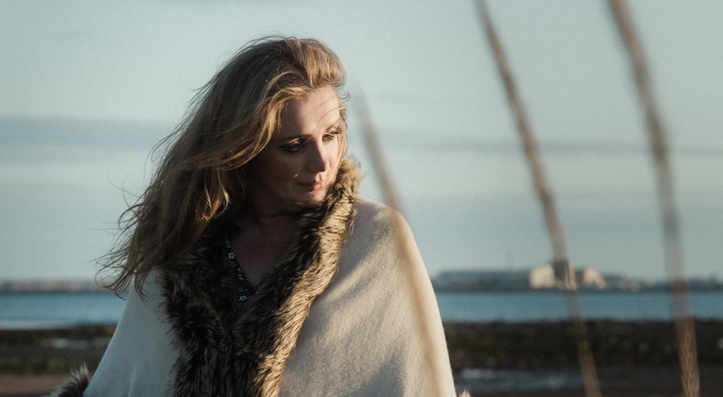 Christine Bovill stands on a beach wearing a faux fur coat