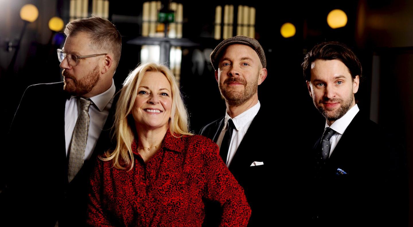 Claire Martin is a blonde woman wearing a red dress, surrounded by three men in black suits, the jazz trio