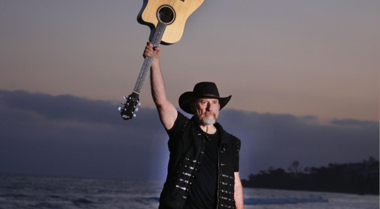 Colin Hay, holding his guitar above his head.