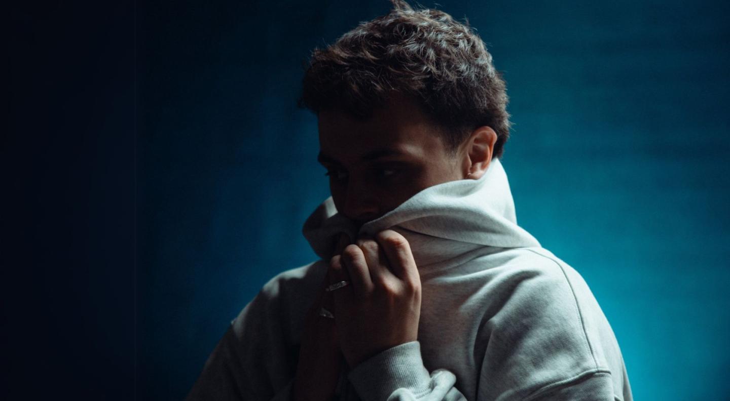 A white male wearing a white hoodie which he has pulled up and is holding over his chin and mouth, standing in front of a white background