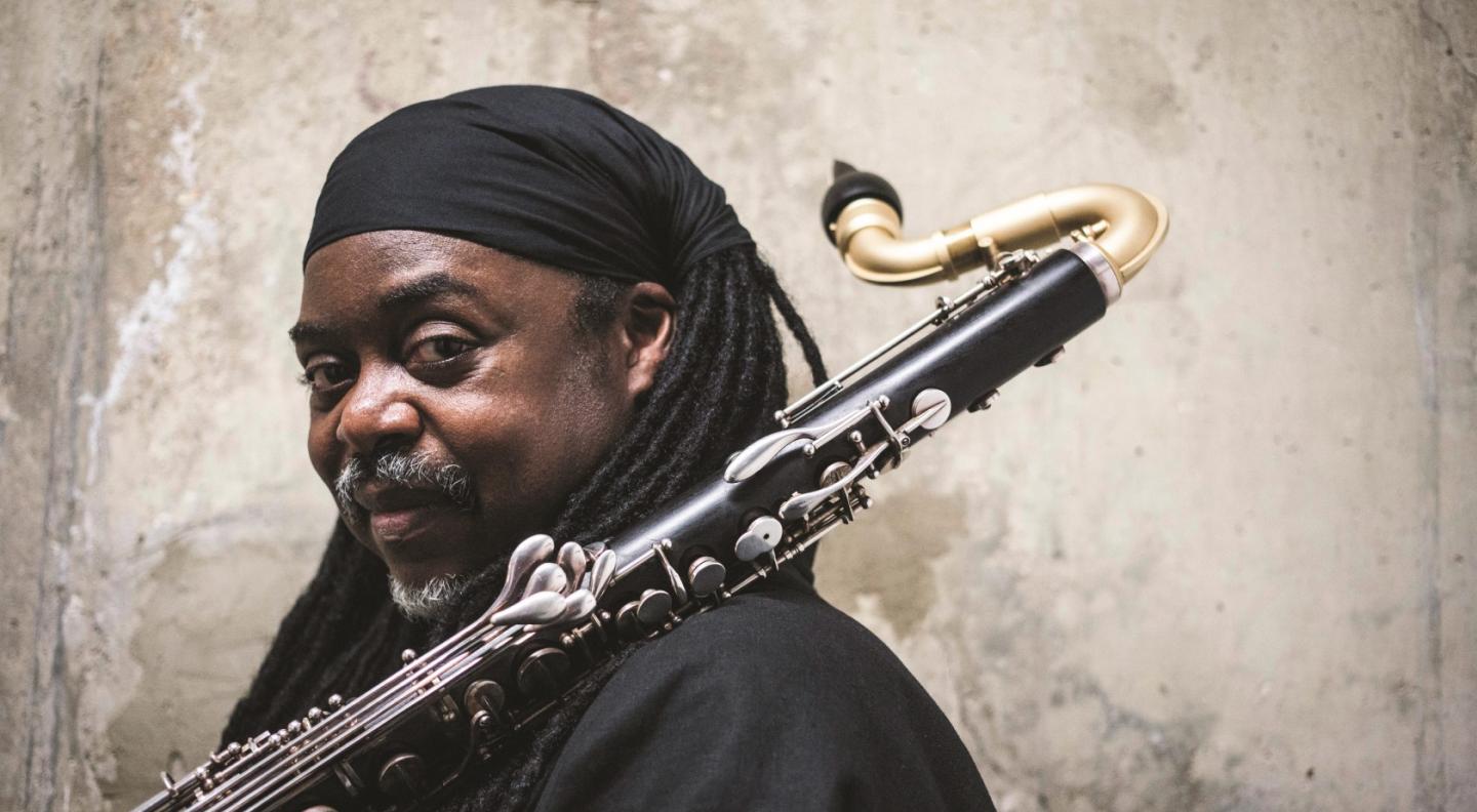 Courtney Pine is dressed in black, saxophone resting on his shoulder, glances to the camera