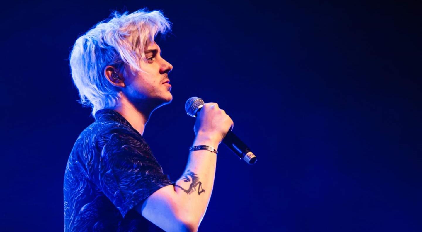 A side-on mid shot of Ethan Mark Nestor-Darling against a blue background. He has bleached blonde hair and a tattoo is visible on the arm in which he holds a microphone up to his mouth