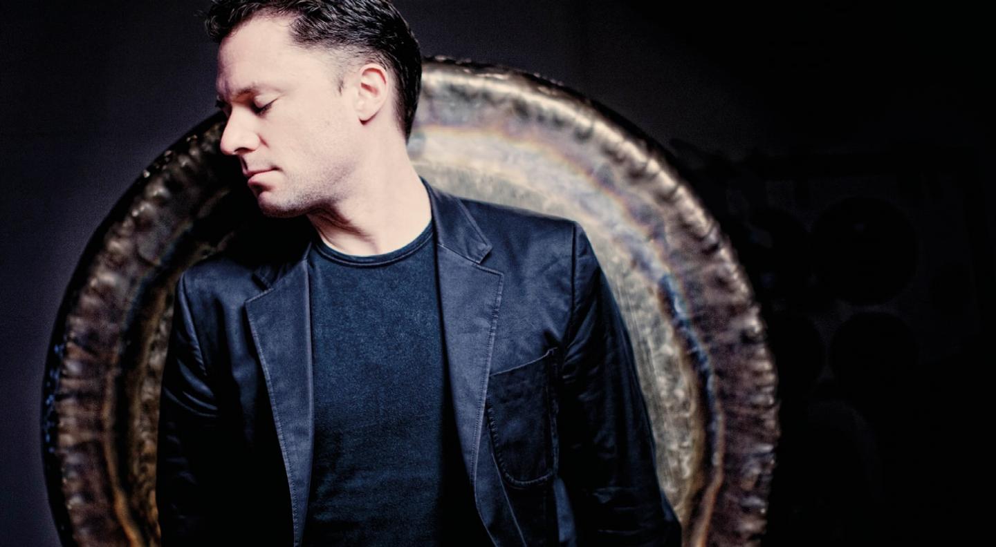 A mid shot of Colin Currie standing with his back against a large gong. He has his eyes closed with his face turned to the side and faced down