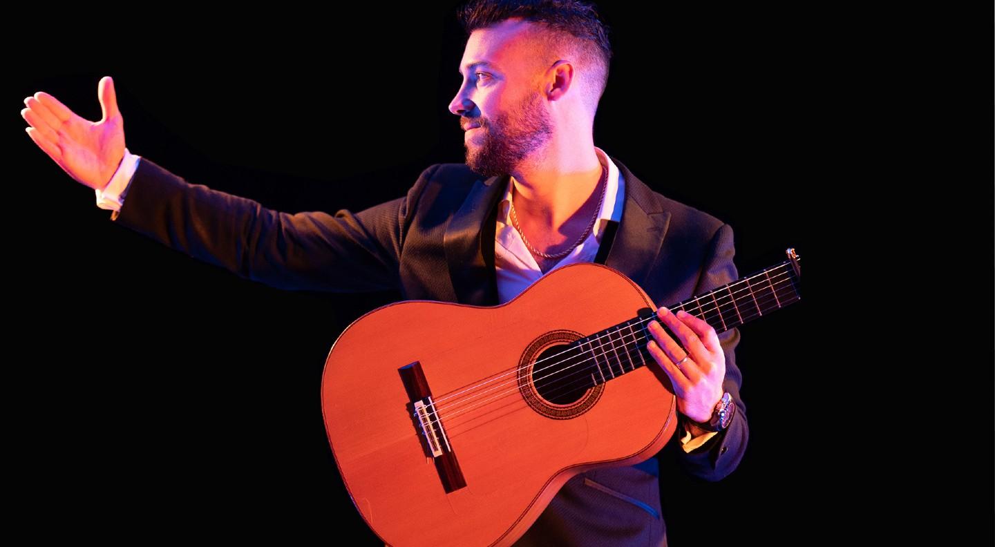 A mid shot in red tones of Daniel Martinez holding his guitar and extending his right arm out to the side as if asking an audience to applaud someone on stage with him