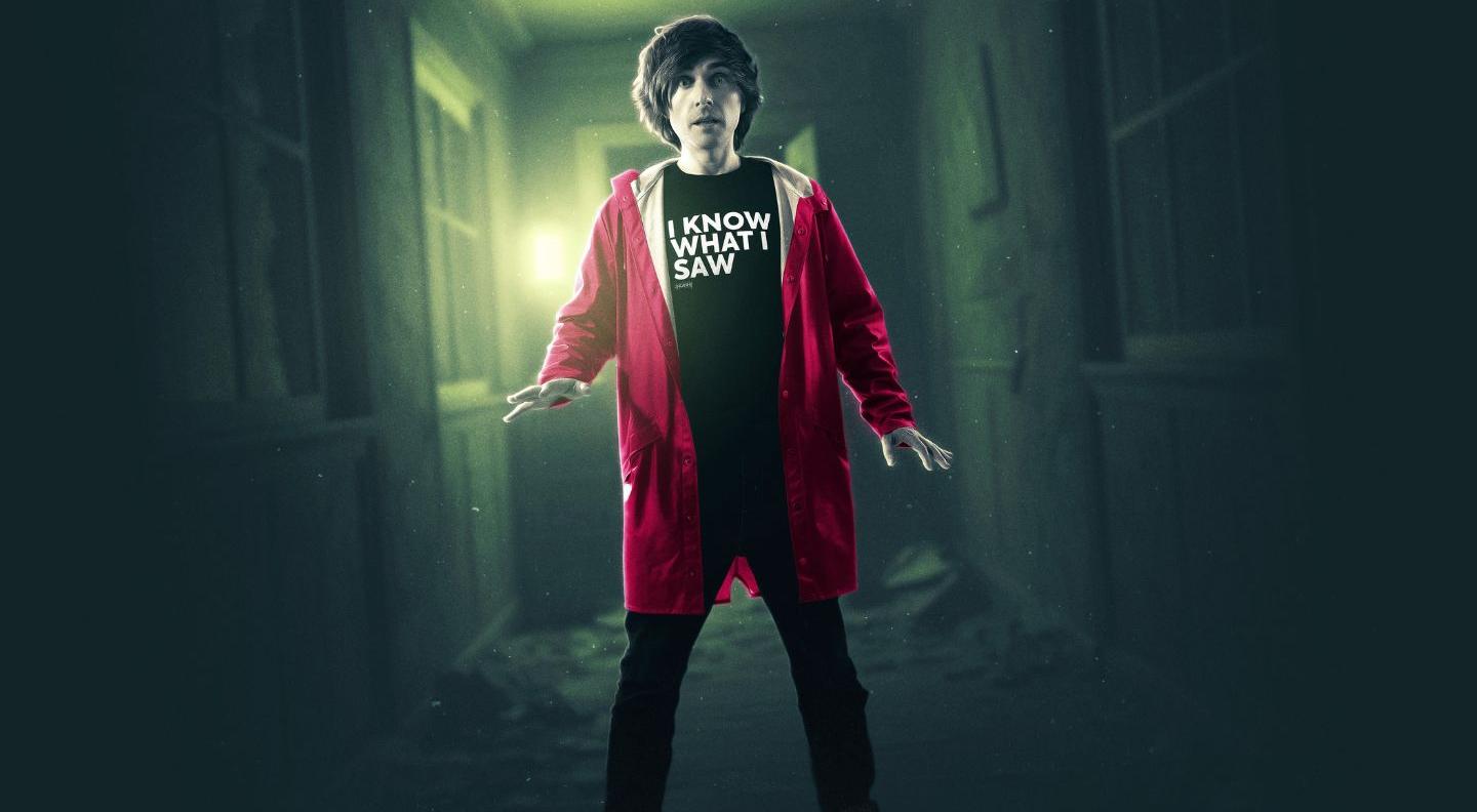 Danny stands in a dark corridor with lit in green, wearing a red coat