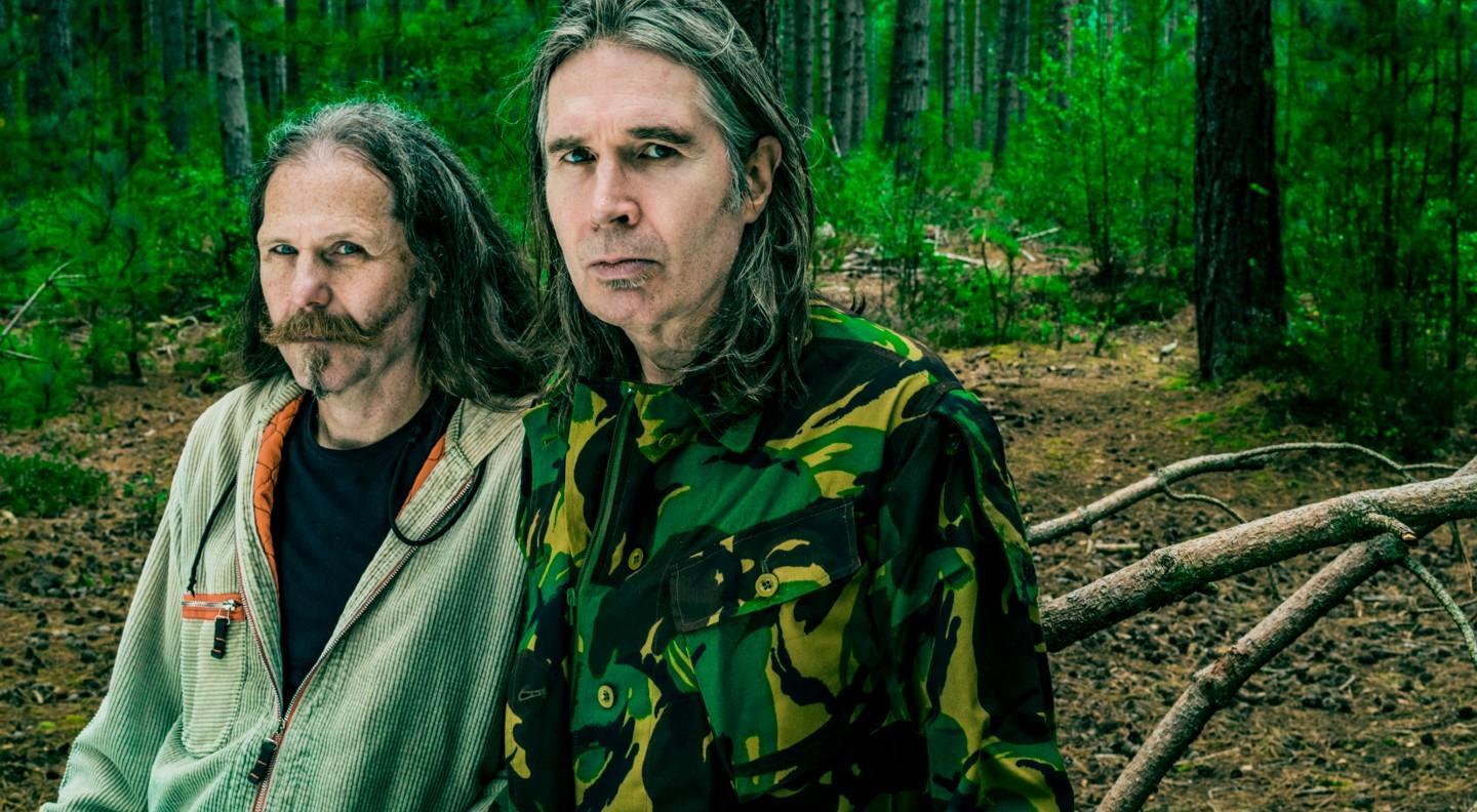 A mid shot of Justin Currie and Iain Harvie in a forest clearing looking out at the camera seriously. There are tree trunks, leaves and roots in the background