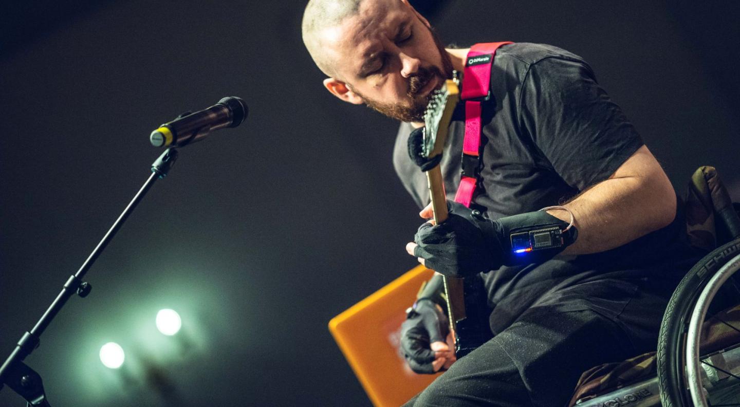 A white man in a wheelchair with a closely shaved head and wearing black leather gloves plays guitar
