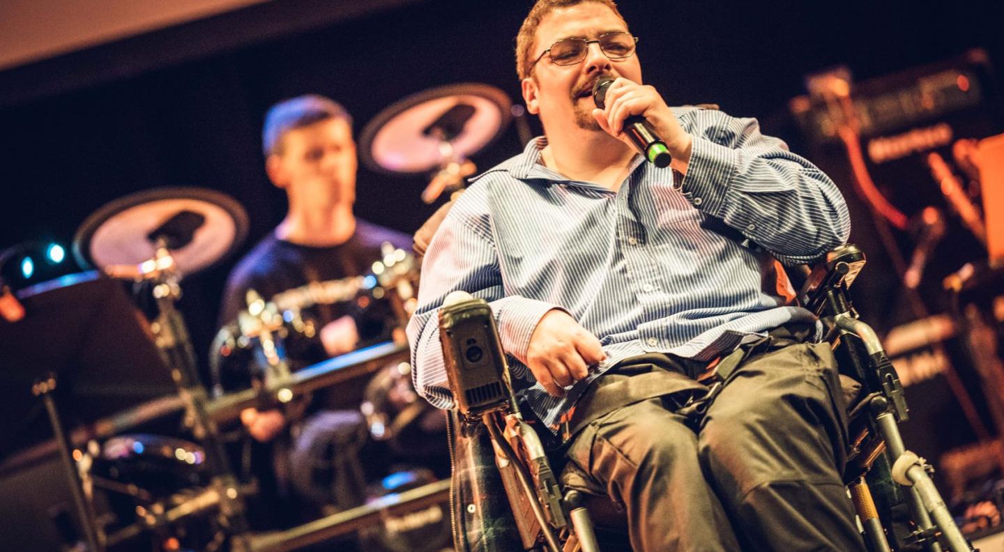 A young white man in a wheelchair sings in to a microphone. Behind him another young white man plays the drums
