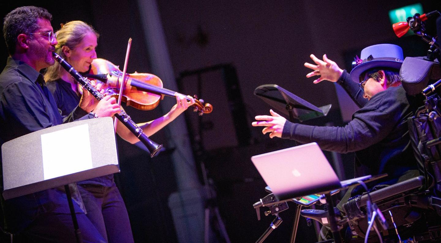 an image of the Drake Scotland digital orchestra.  A man in an electric wheelchair conducts a violin player 
