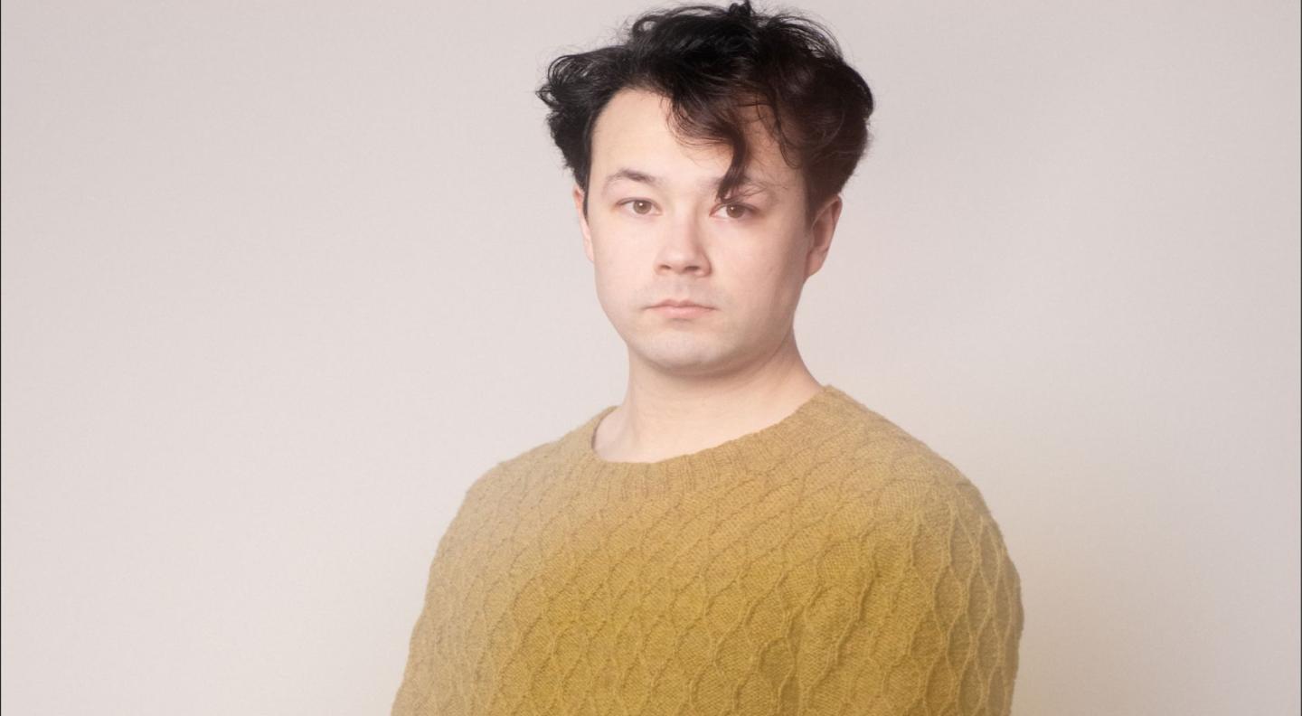 Sean Shibe wearing a mustard colour jumper standing in front of a white background