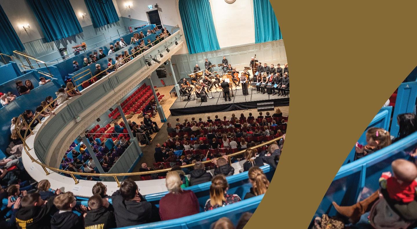 A view down to The Queen's Hall stage from the balcony showing an auditorium full of people. A gold swoosh has been placed over the top