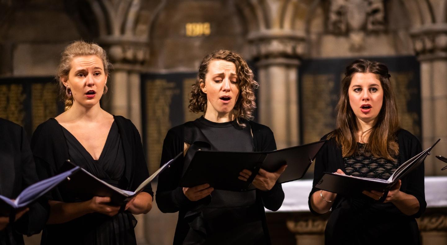 Three female members of Dunedin' a cappella consort perform in a church surrounding all wearing black and holidng music folders. Image credit Andy Catlin