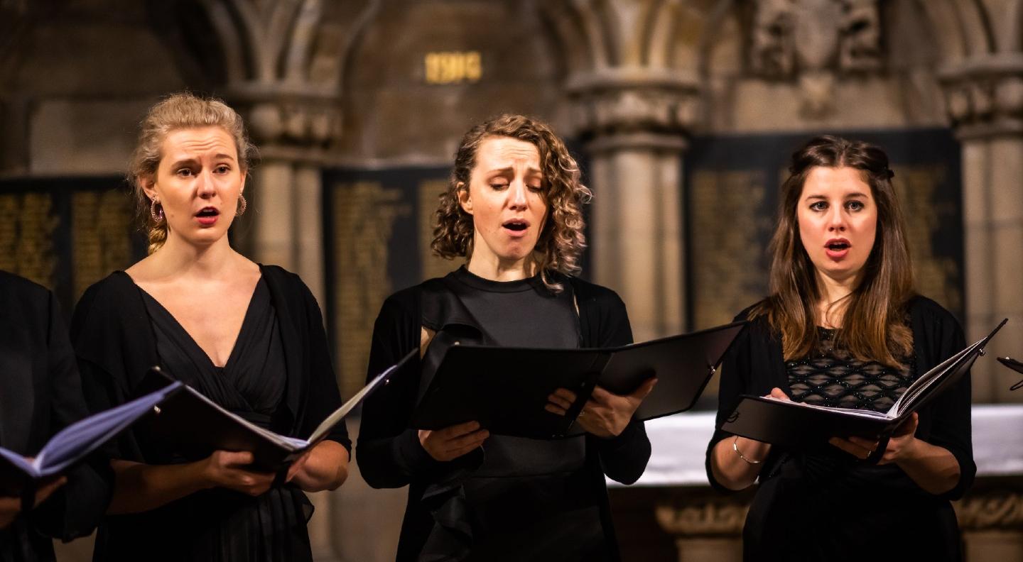 Three female members of Dunedin' a cappella consort perform in a church surrounding all wearing black and holidng music folders. Image credit Andy Catlin