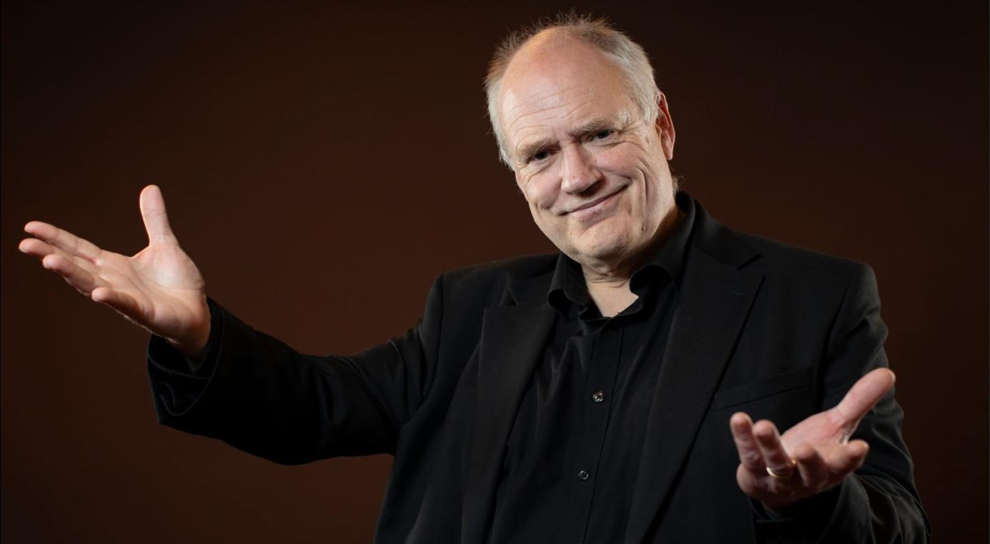 John Butt posing with his arms open in front of a brown background