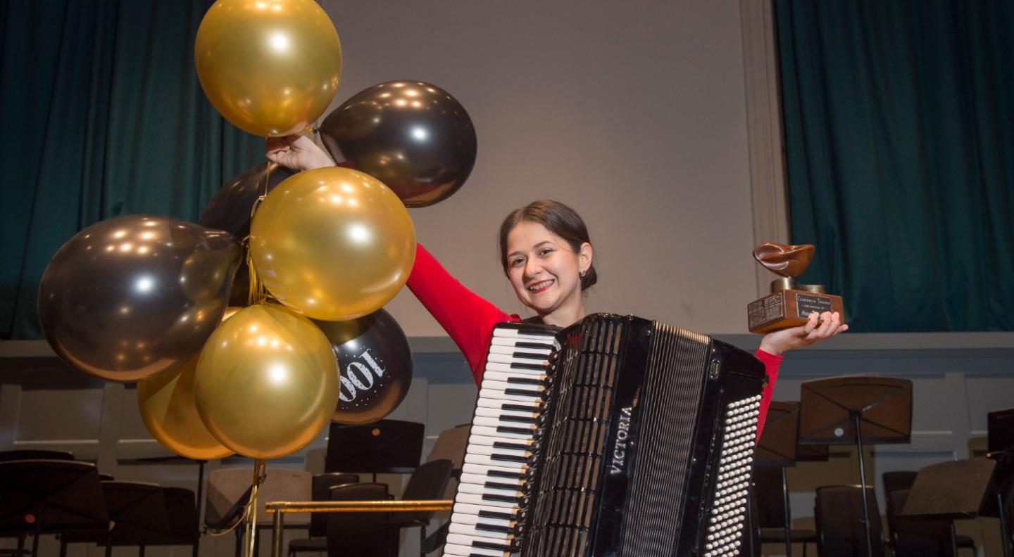 An image of a woman wearing a red top with an accordion slung in front of her. She is holding gold and black balloons in one hand and a trophy in th eother. 