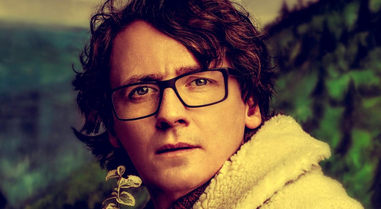 Ed Byrne stands in front of a mountainous backdrop wearing a sheepskin collar coat. He looks out at the camera pensively