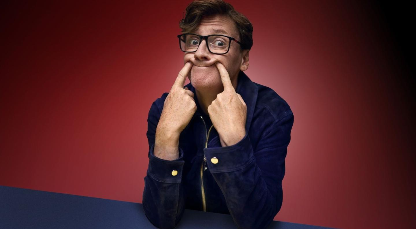 Ed Byrne sitting at a yellow table, pushing his mouth into a smile with his pointer fingers