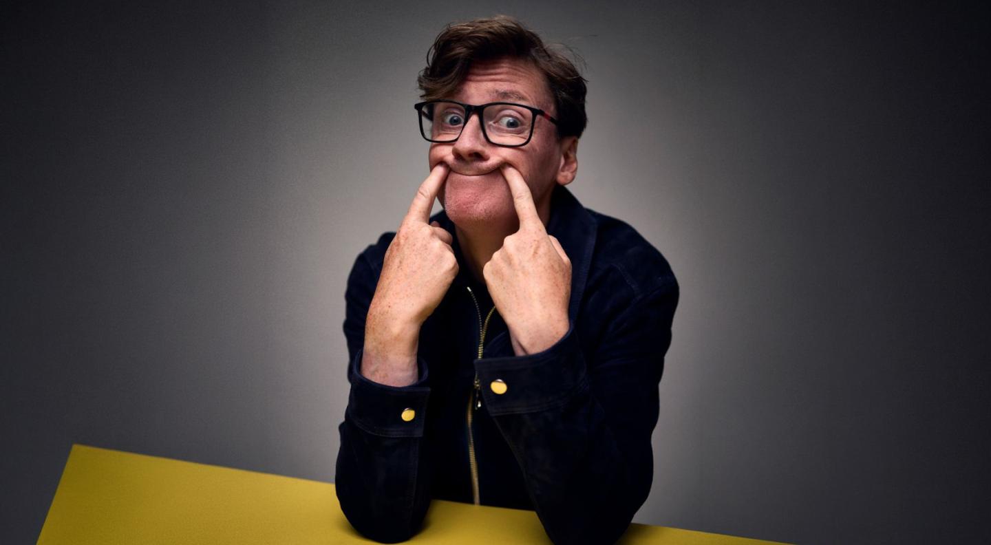 Ed Byrne sitting at a yellow table, pushing his mouth into a smile with his pointer fingers