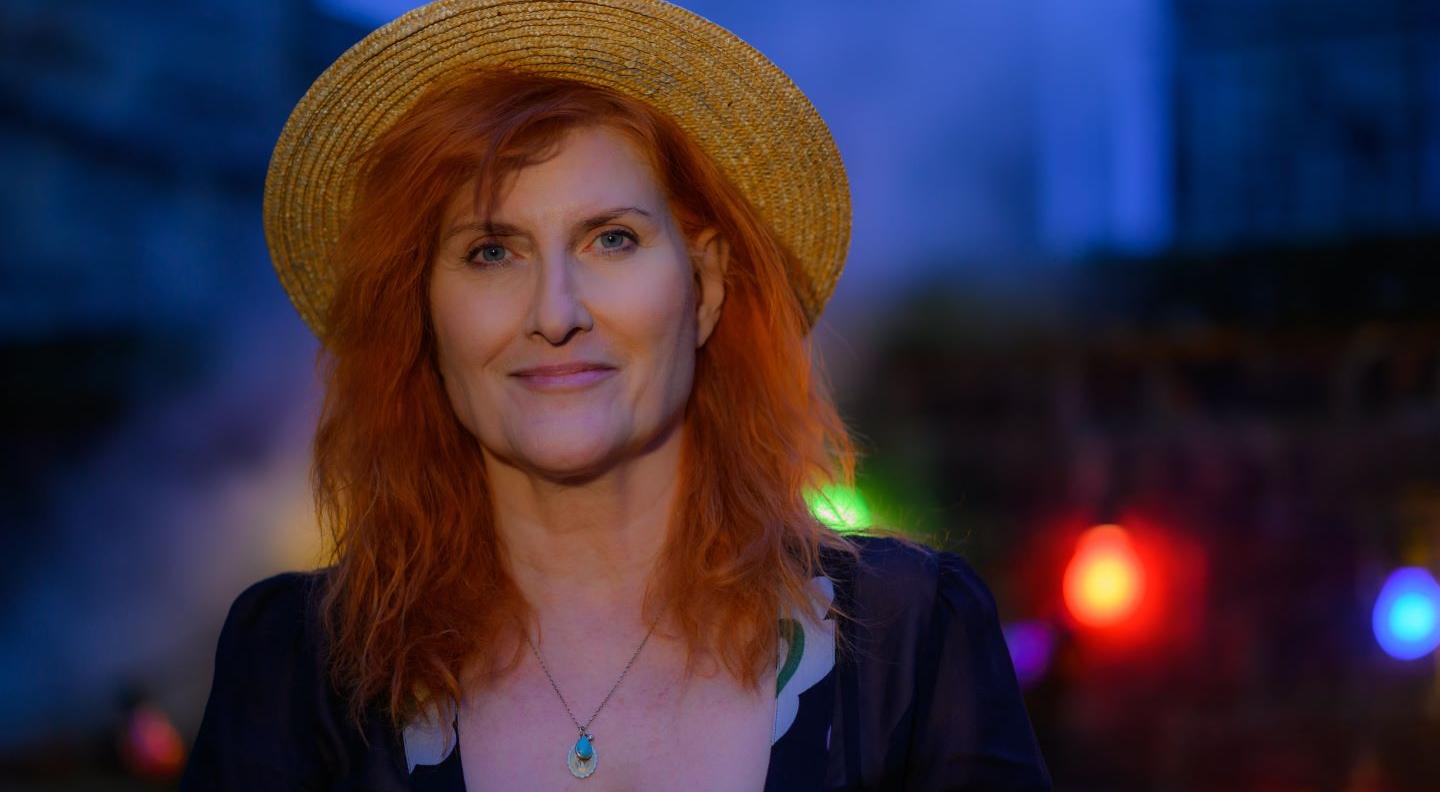 A full colour mid shot of Eddi Reader taken against a blurred cityscape at nightime. She wears a hat on the back of her head and smiles out at the camera. Copyright Sean Purser