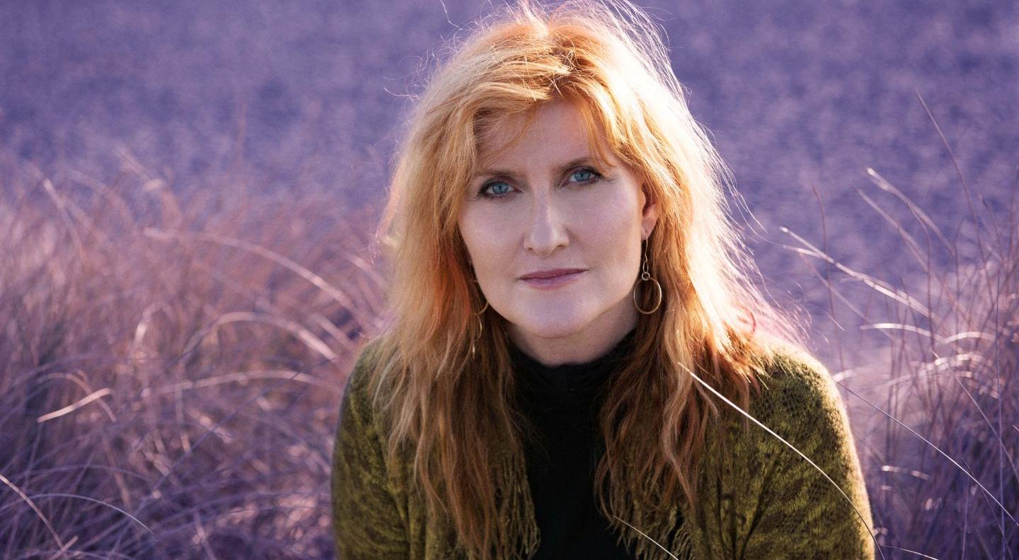A white woman with red hair and blue eyes sits in a field of purple-tinted grass, wearing a brown turtleneck jumper