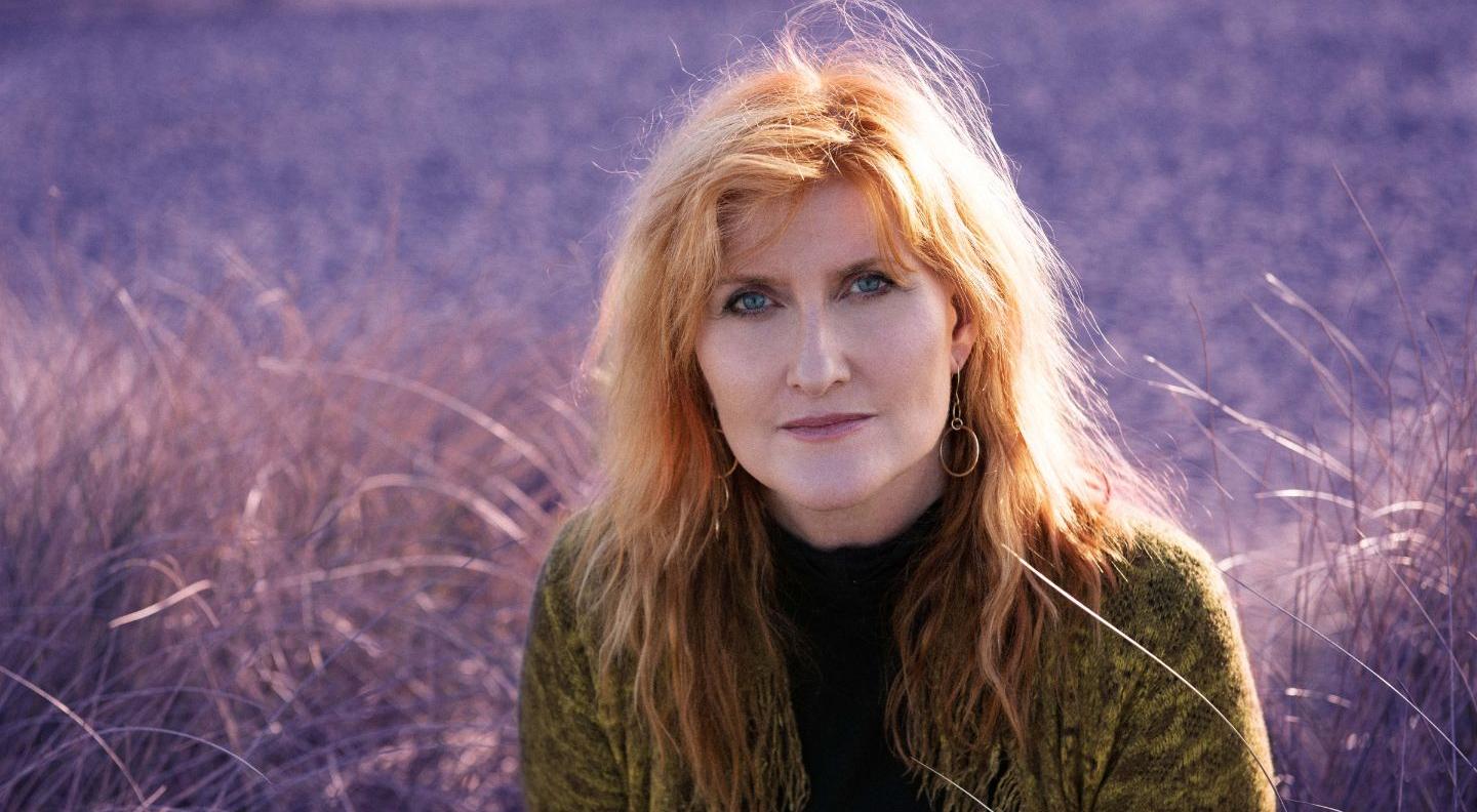 A white woman with red hair and blue eyes sits in a field of purple-tinted grass, wearing a brown turtleneck jumper