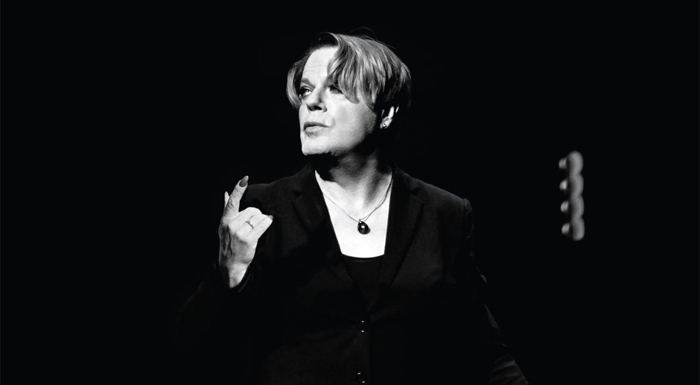 Eddie Izzard posing in front of a plain black backdrop, in black and white.
