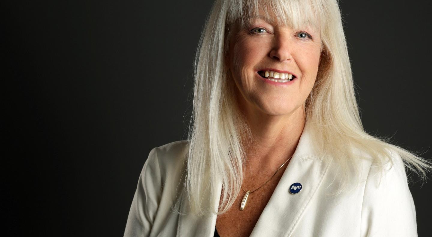 A mid shot of Lesley Riddoch standing against a black background smiling at the viewer