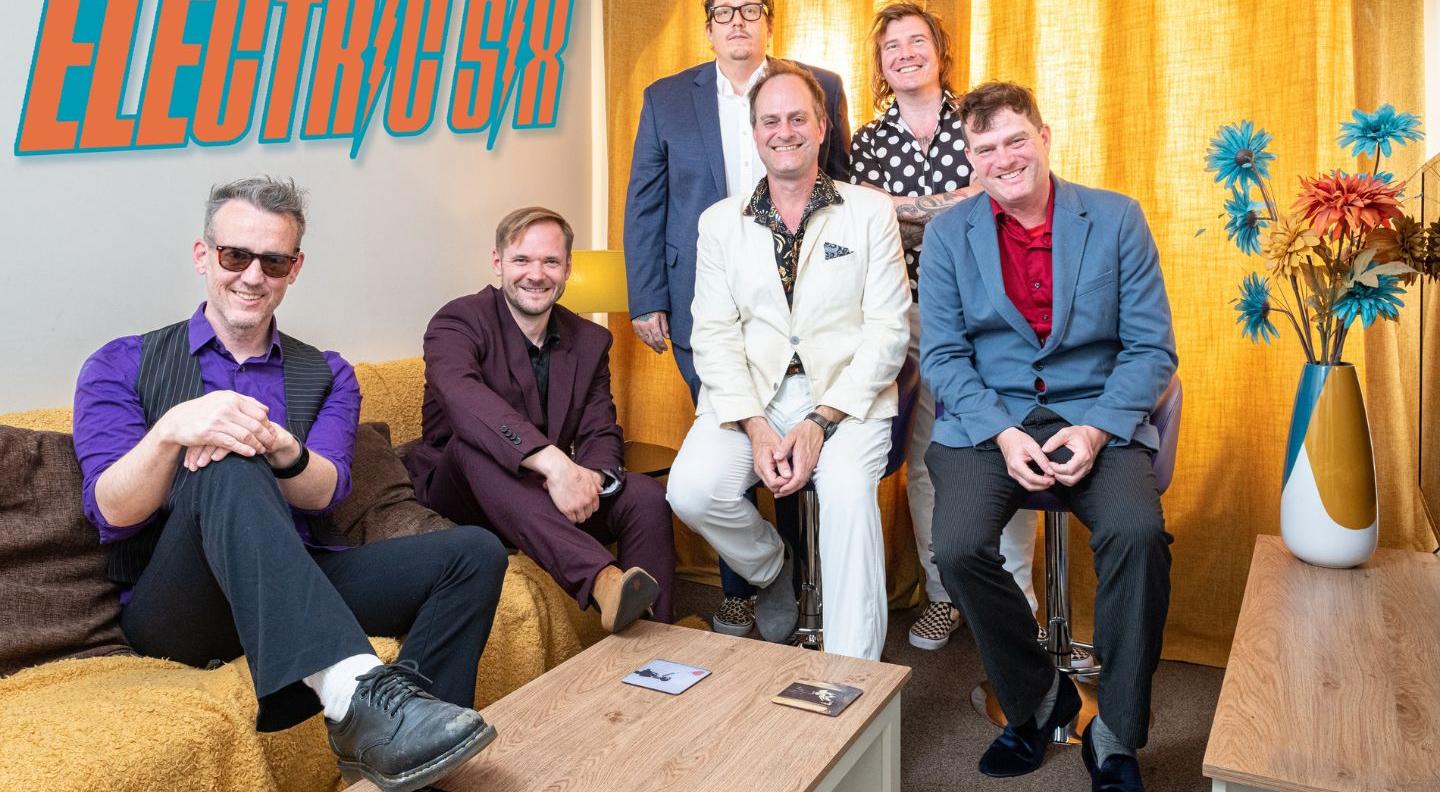 An image of the band Electric Six, six white men sat on a sofa around a coffee table. There's a sign on the wall which says 'Electric Six'