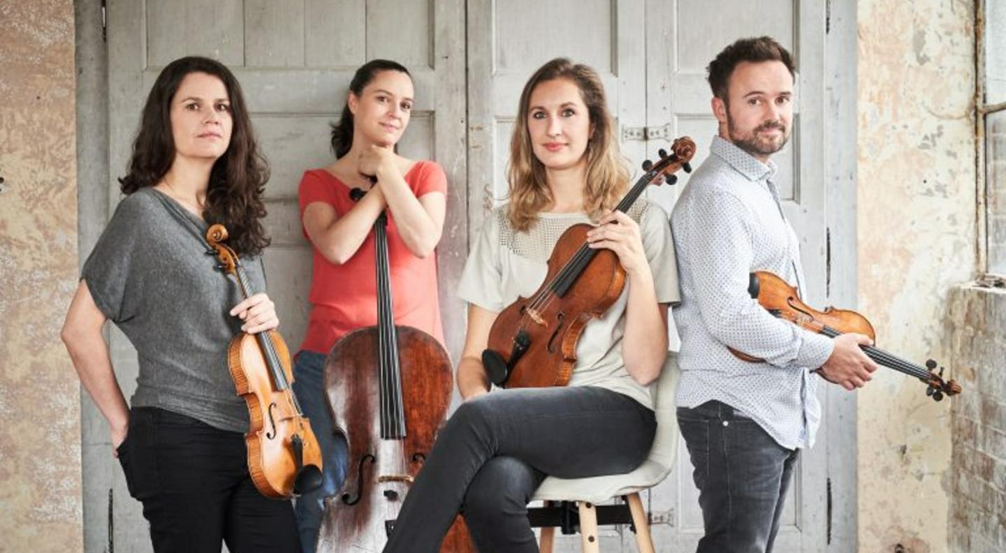 The Elias Quartet - three white women and one white man - pose seated and standing with their stringinstruments in front of a white panelled wall