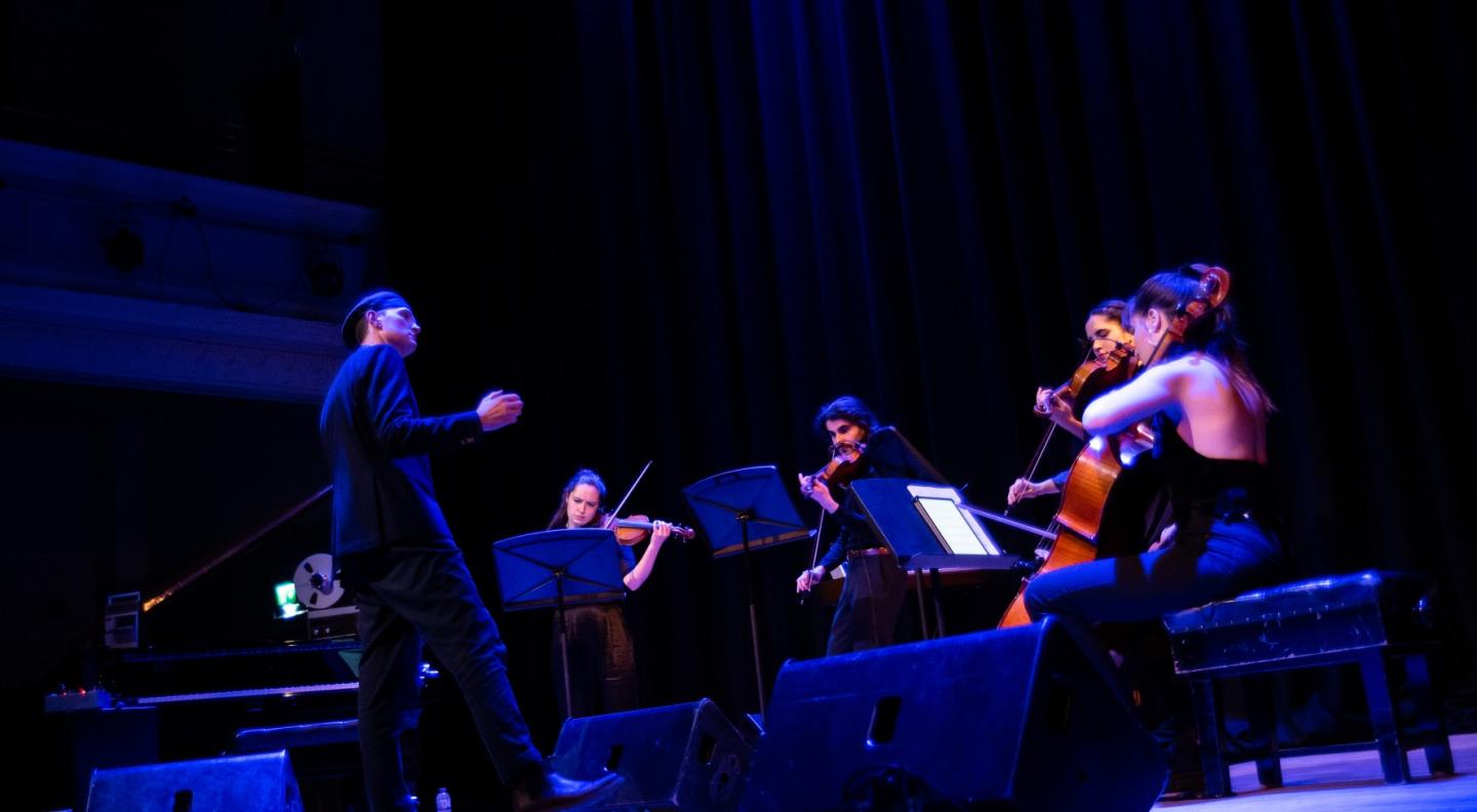 A tall white man wearing a cap stands by a grand piano conducts a string quartet