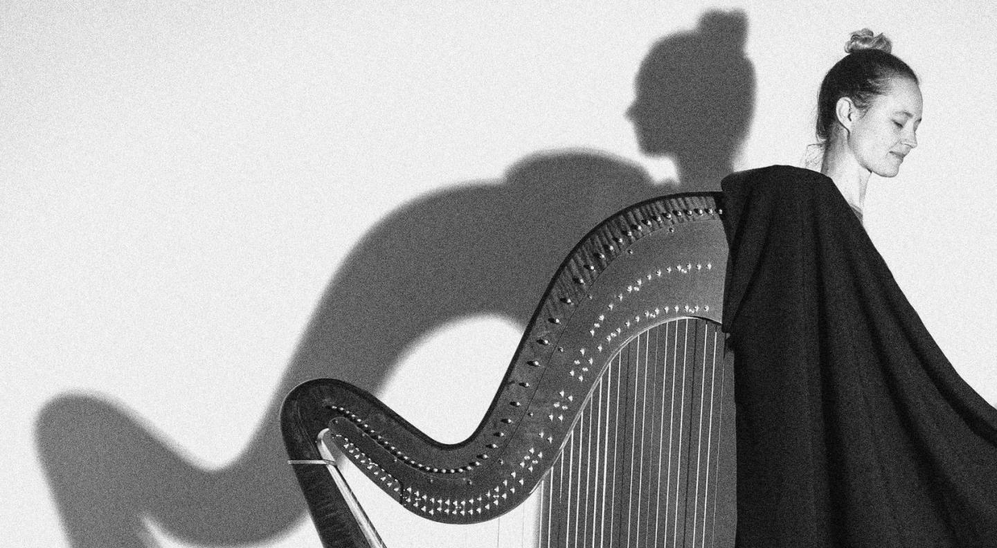 A black and white image of a white woman standing with her back to a harp in a dress which mirros the harp shape. Shadows are reflected on the blank wall behind