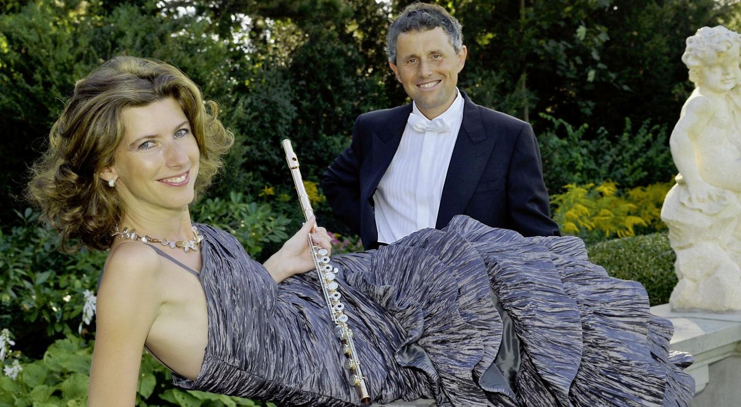 Eva Oertle and Vesselin Stanev sit in a garden. He wears a black tuxedo and she reclines in a grey ballgown, holding her flute