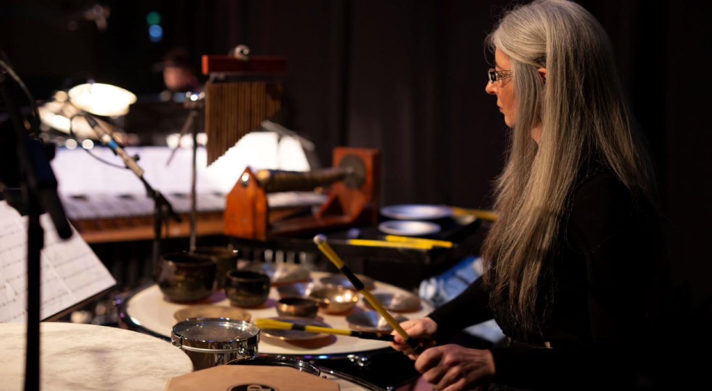 A white woman with long silver hair and glasses holds drum sticks in her hands and has various percussion instruments around her on stage