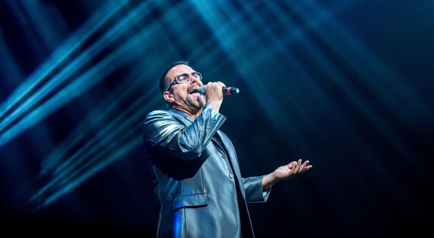 A man dressed as George Michael sings into a microphone on  a dark stage. He wears a leather blazer and sunglasses