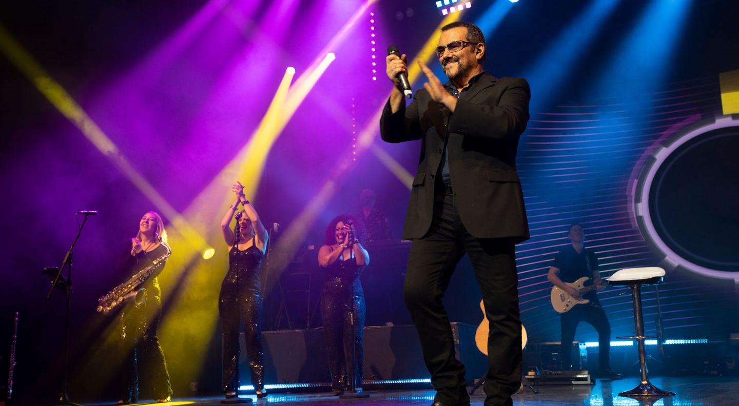 an image of the artist on stage, dressed as George Michael in a suit and singlasses, with backing singers  and musicians