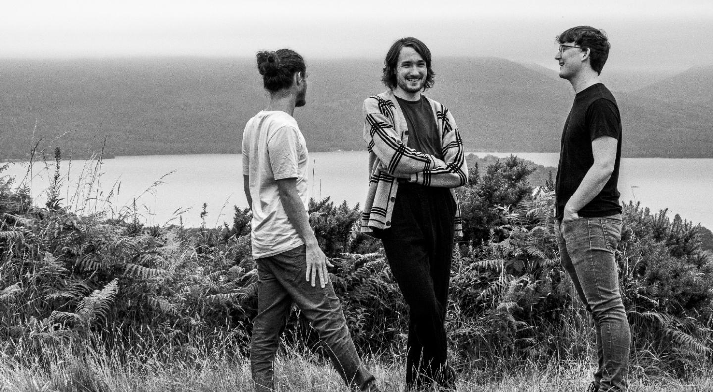 A black and white full length shot of the Fergus McCreadie Trio standing in an outdoor setting with a loch, hills and a stone wall beind them