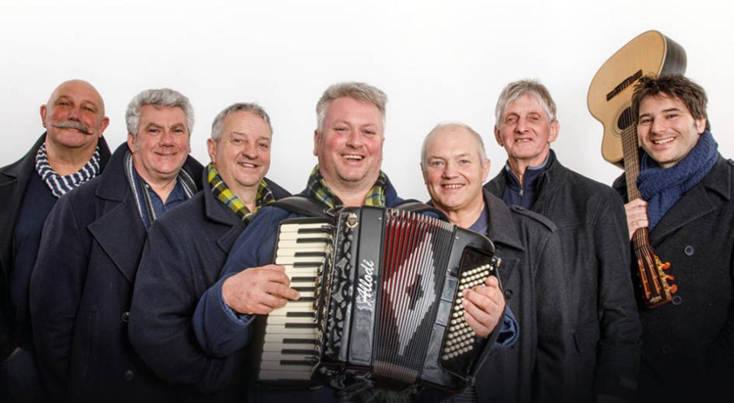 The Fisherman's Friends, all smiling, wearing dark coats and different coloured scarves stand in a line looking out at the camera. Jason is in the middle holding his accordion. Toby on the far right has his guitar over his shoulder.